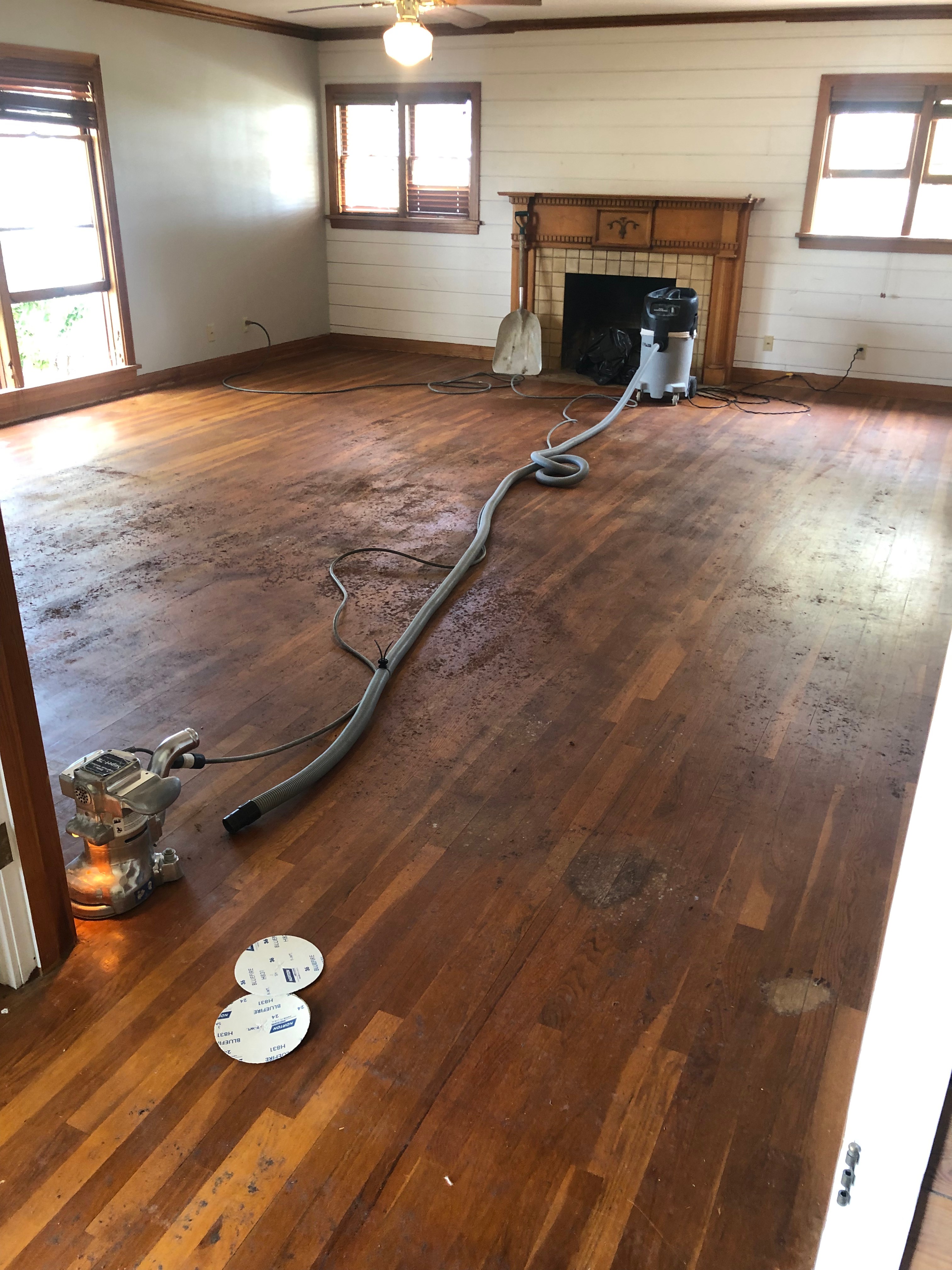 Before restoration: Heavily stained and worn hardwood floor with dark discoloration, professional sanding equipment visible in living room